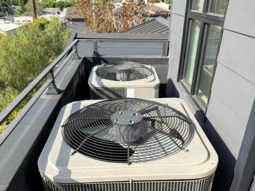 Two air conditioner condensers installed on a narrow rooftop deck beside a home's exterior wall